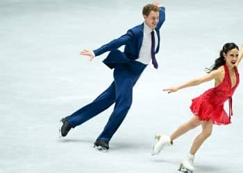 Madison Chock and Evan Bates skate parallel to each other at NHK Trophy (she on the right, he on the left). She wears a bright red halter dress while he wears a blue suit with a white shirt. They both smile, their arms spread wide as they skate.